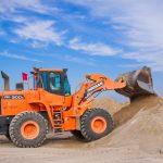 An orange bulldozer at work, moving sand under a clear blue sky on a construction site.